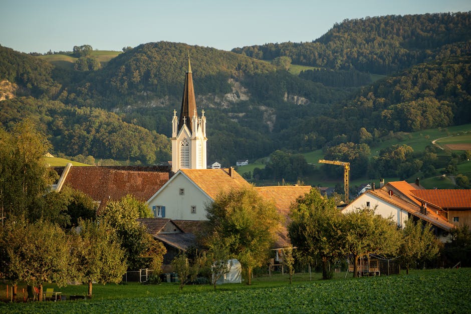 Quaint village church with spire surrounded by lush green hills and trees, bathed in daylight.